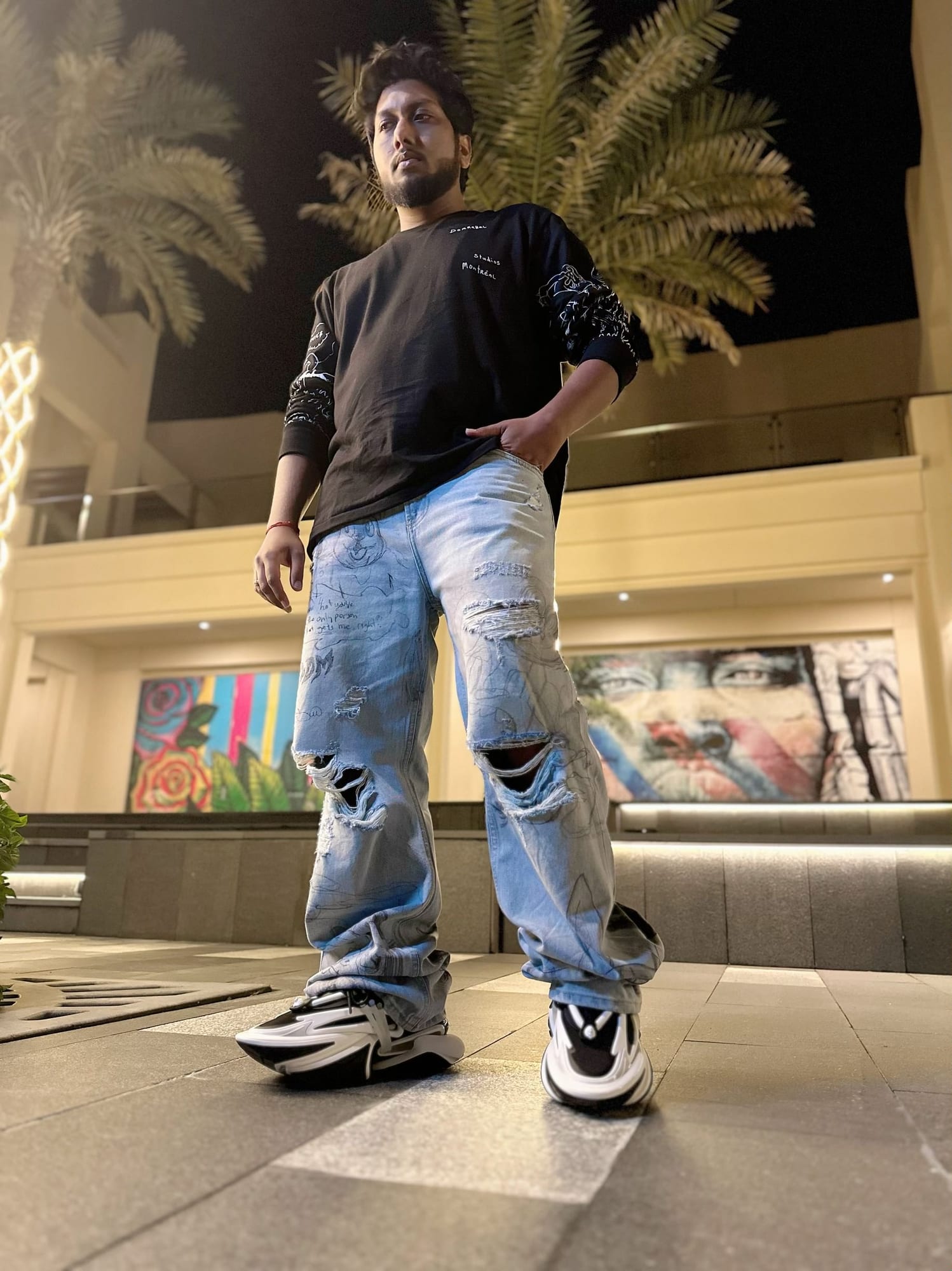 Vinay Anuragh photographed from below at night in a black graphic sweatshirt, beneath palm trees and warm architectural light