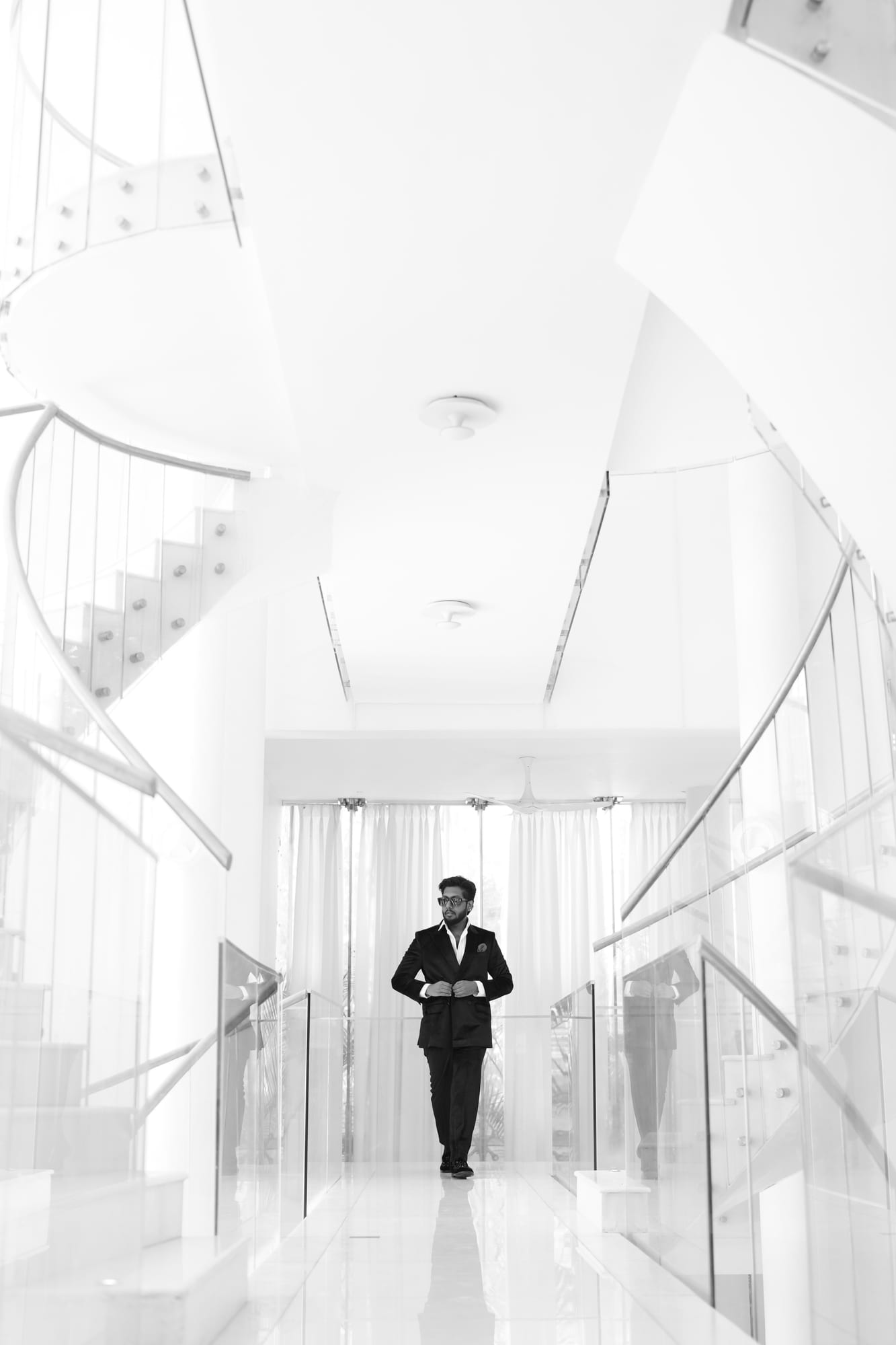 Vinay Anuragh in a black velvet suit, walking down a white glass-railed corridor between two spiral staircases