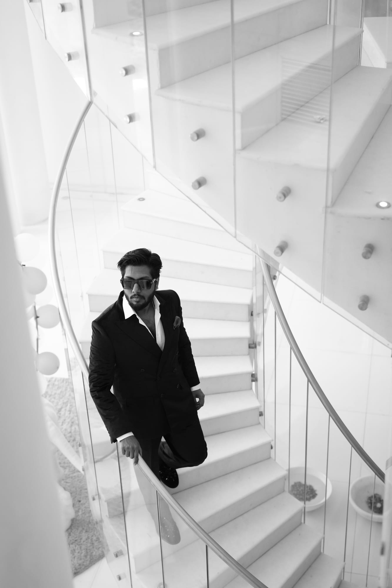 Vinay Anuragh in a black double-breasted suit and dark sunglasses, descending a white spiral staircase